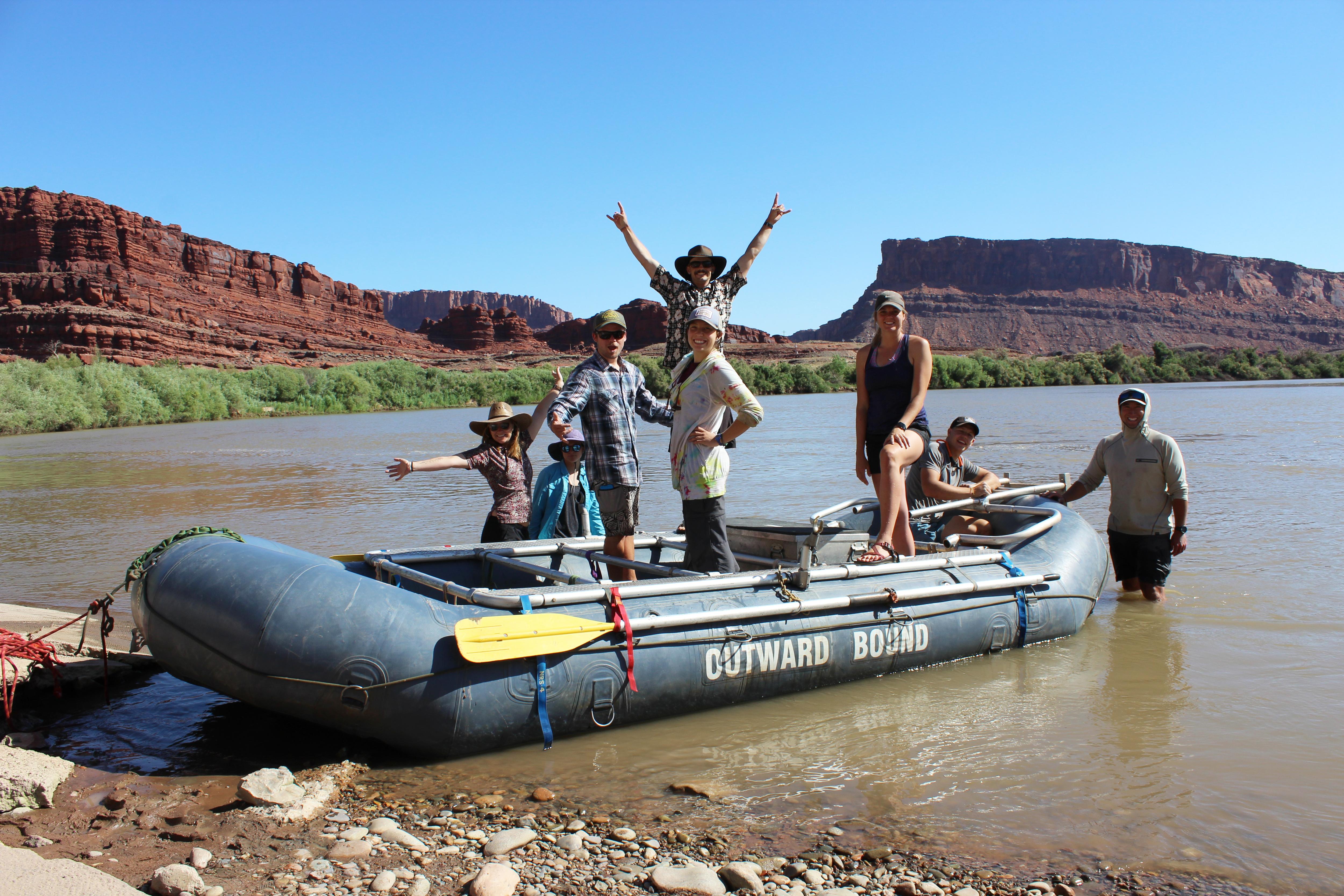 The image shows a group of people on a raft in a river. They are posing for the camera, with some raising their arms in the air. The raft is labeled "COTWARD BOUND". The background features a scenic view of red rock formations and a clear blue sky.